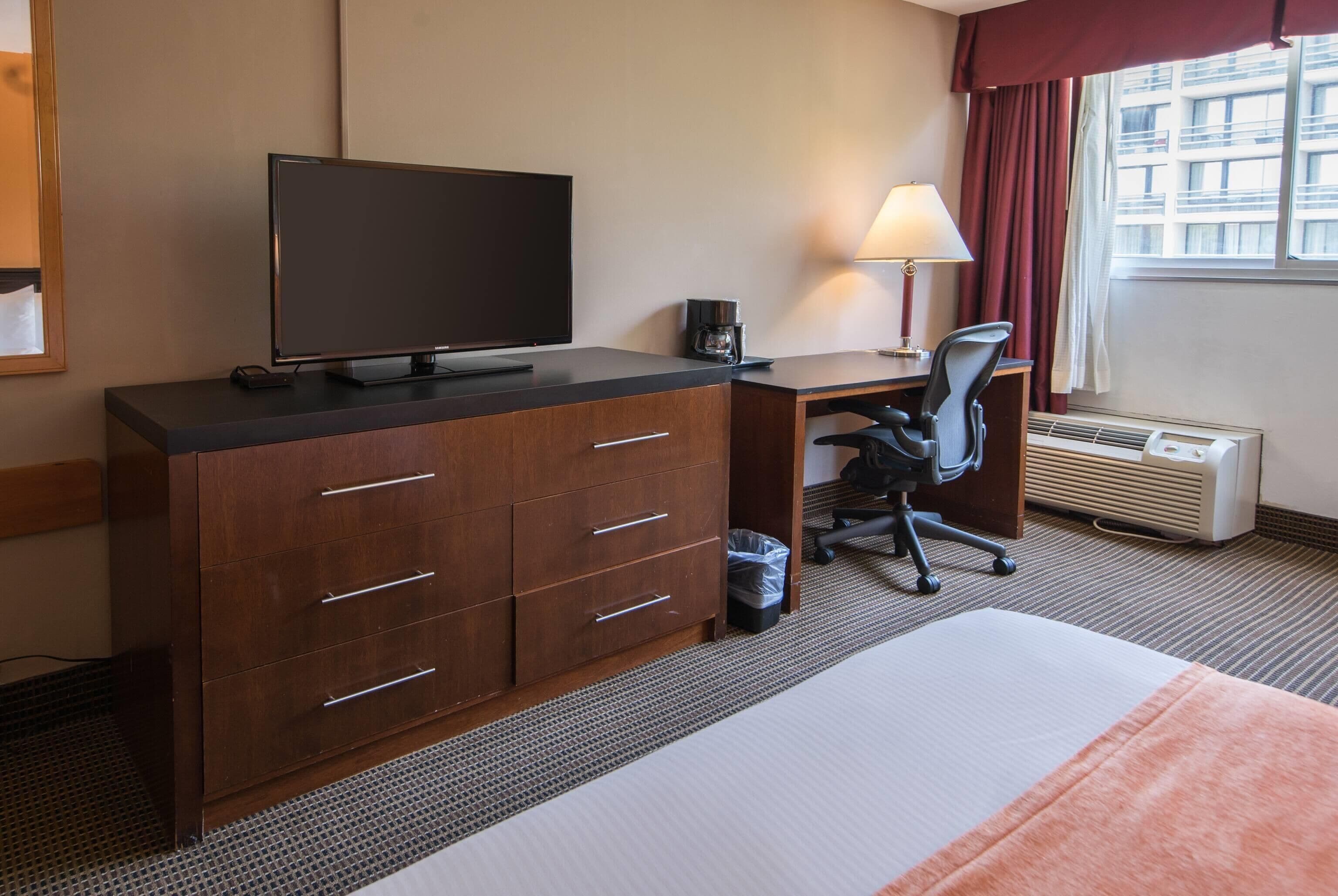 A hotel room with a bed, a desk with a computer, a TV, a lamp, and a window with curtains.