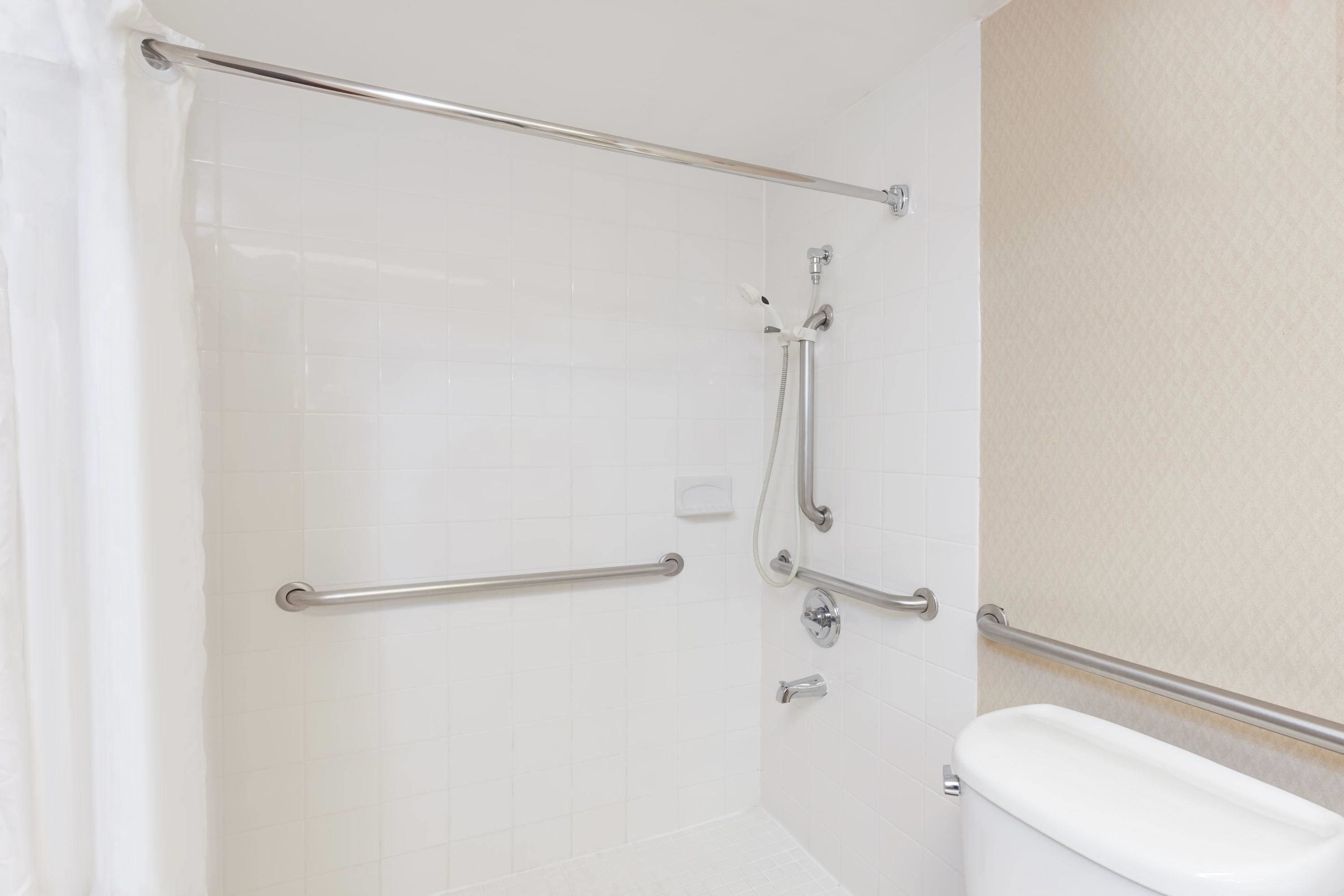 Une salle de bain avec des murs recouverts de carreaux blancs, une douche avec une porte en verre et des toilettes équipées d’une barre d’appui.