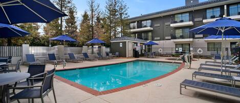 A swimming pool with lounge chairs and umbrellas, a small building, and a multi-story building with balconies.