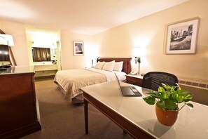 Ein Hotelzimmer mit einem Bett, einem Schreibtisch mit Laptop, einem Sessel, einer Topfpflanze und einem gerahmten Bild an der Wand.