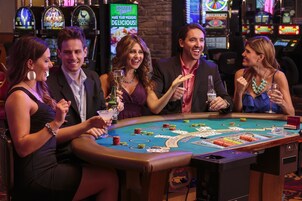 A group of people seated around a casino table with chips and cards, with slot machines in the background.