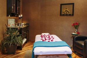 A massage room with a massage table, a potted plant, a small table with candles, and a chair.