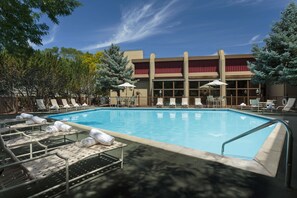 A swimming pool with lounge chairs and umbrellas, surrounded by trees and a building with large windows.