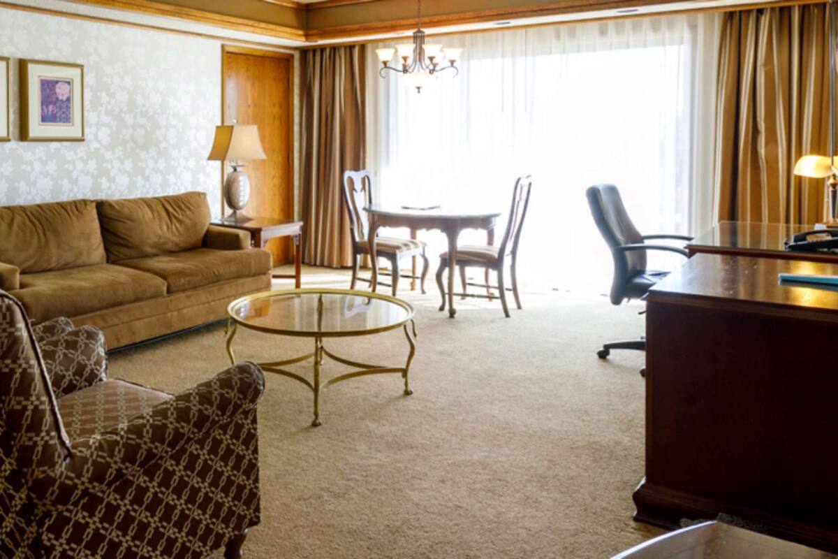 A hotel room with a sofa, armchairs, a dining table with chairs, a desk, and lamps.