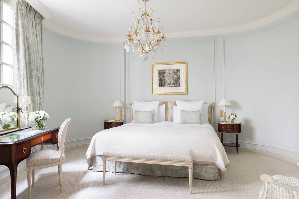 A bedroom with a large bed, a desk with a chair, a chandelier, and a framed picture on the wall.