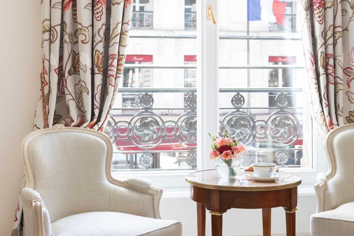Une pièce comprenant deux fauteuils rembourrés, une petite table en bois avec une théière et une tasse, et une fenêtre agrémentée de rideaux décoratifs.