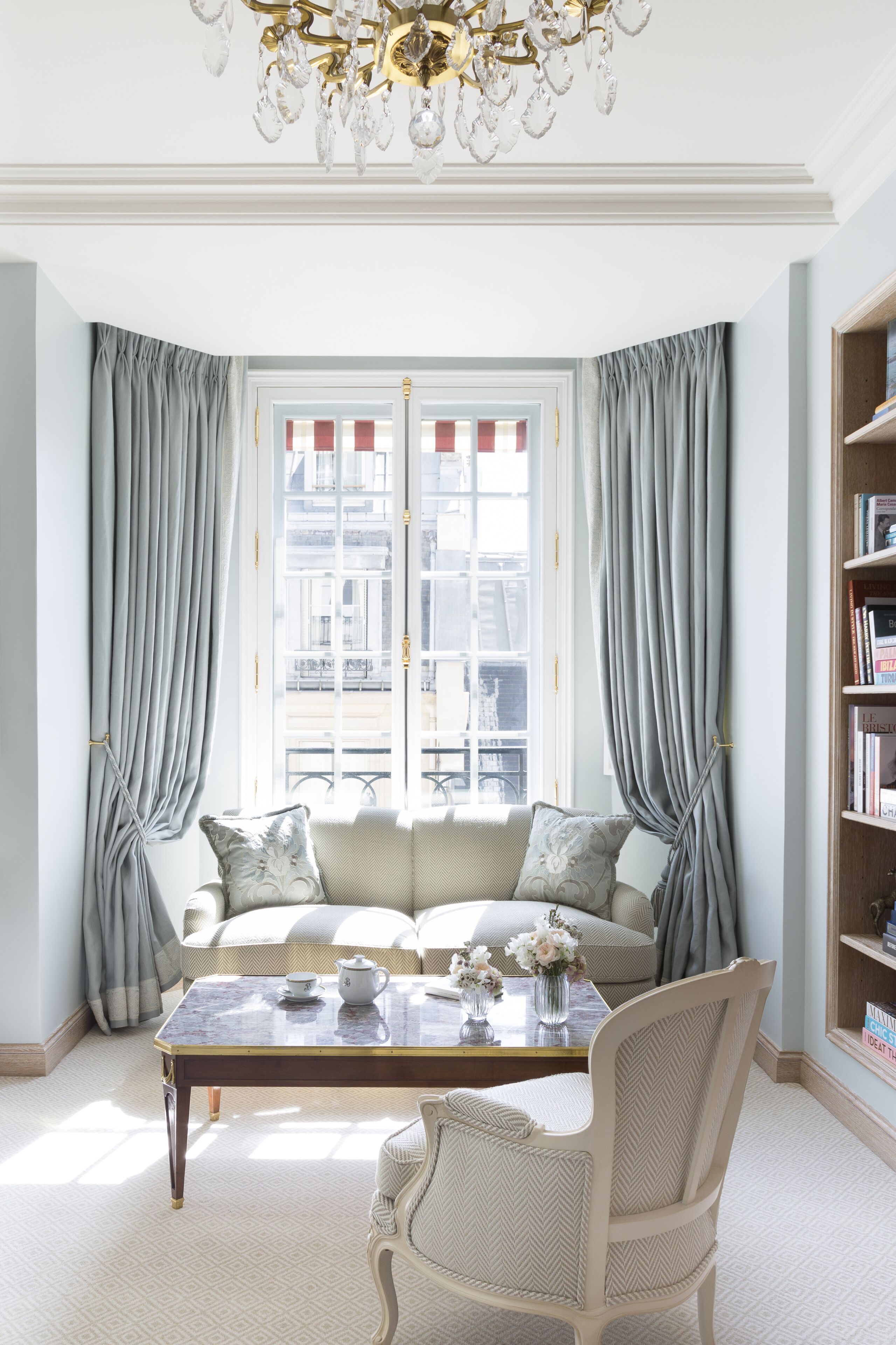 A room with a sofa, a table, and a chair. There is a window with curtains and a chandelier.