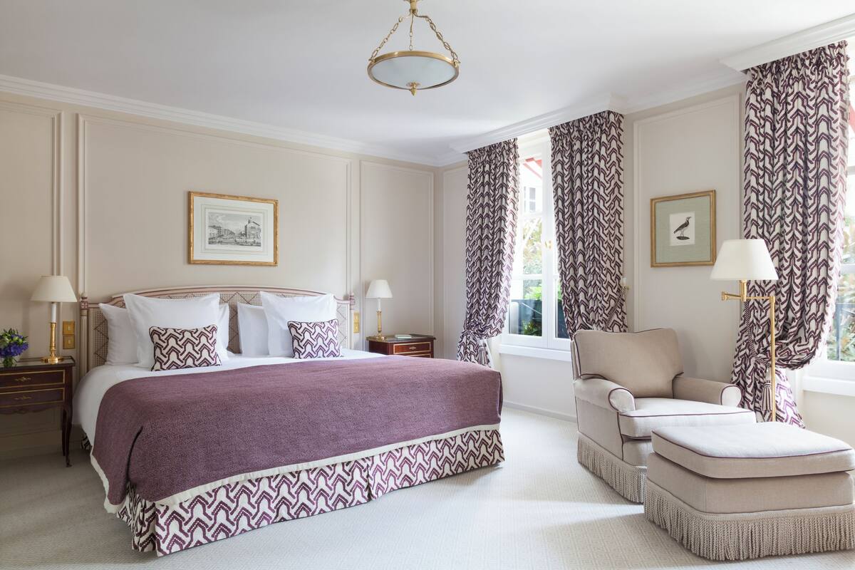 A well-appointed bedroom with a large bed, a chaise lounge, and a small ottoman. There are two bedside tables with lamps, a framed picture on the wall, and patterned curtains.