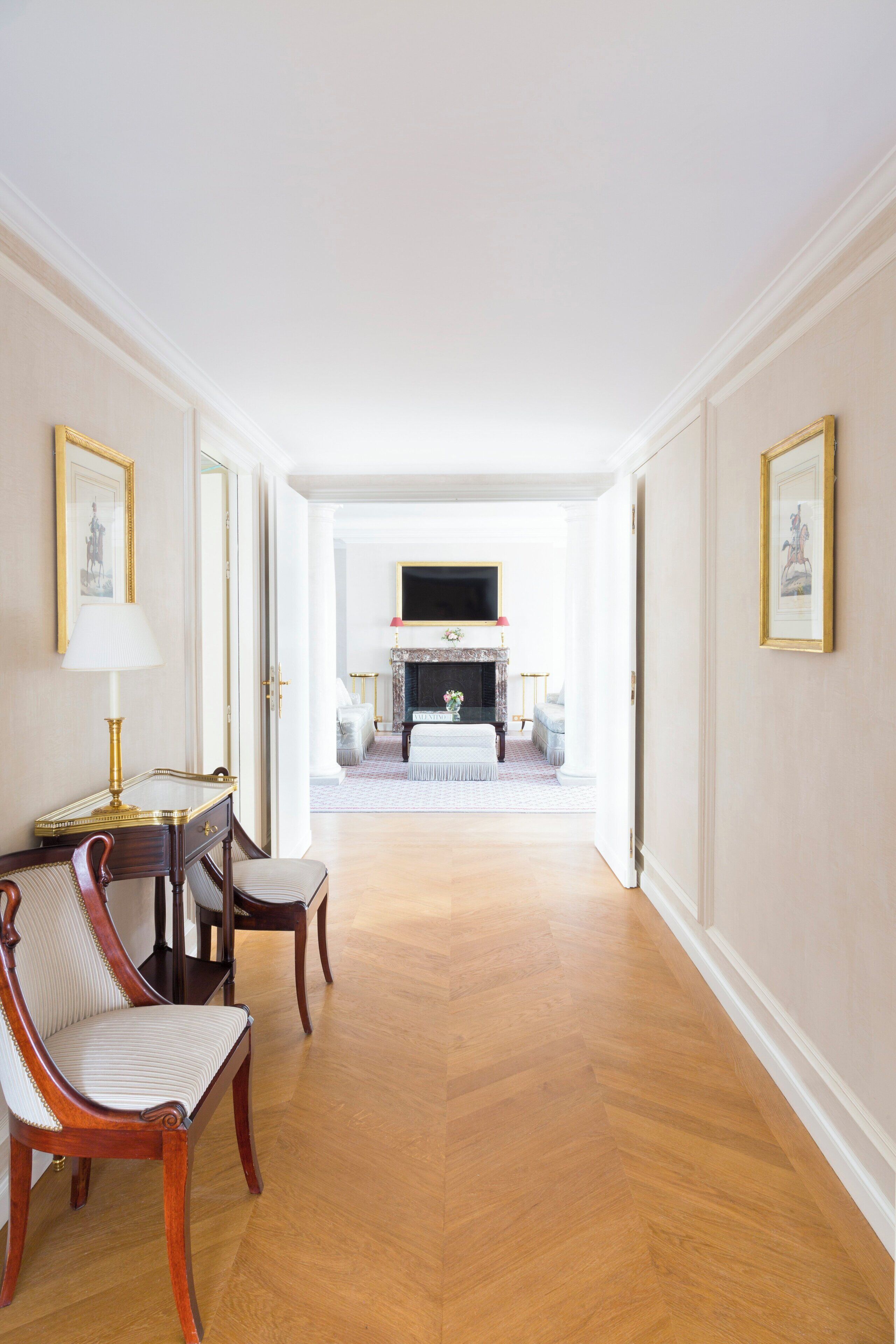 A hallway with a wooden floor leading to a room with a fireplace, framed pictures on the walls, and a desk with a lamp.