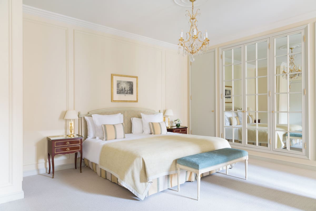 A bedroom with a large bed, bedside tables, a bench, a chandelier, and a large mirror.