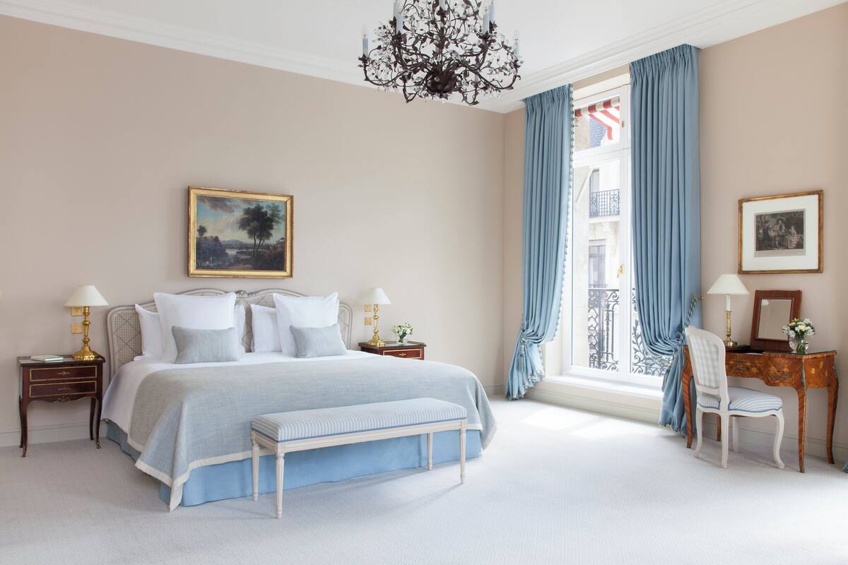 A bedroom with a large bed, a desk, a chair, a chandelier, and a window with curtains.