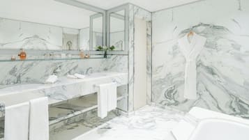 A bathroom with marble walls, a glass shelf with toiletries, a bathtub, and a robe hanging on a hook.