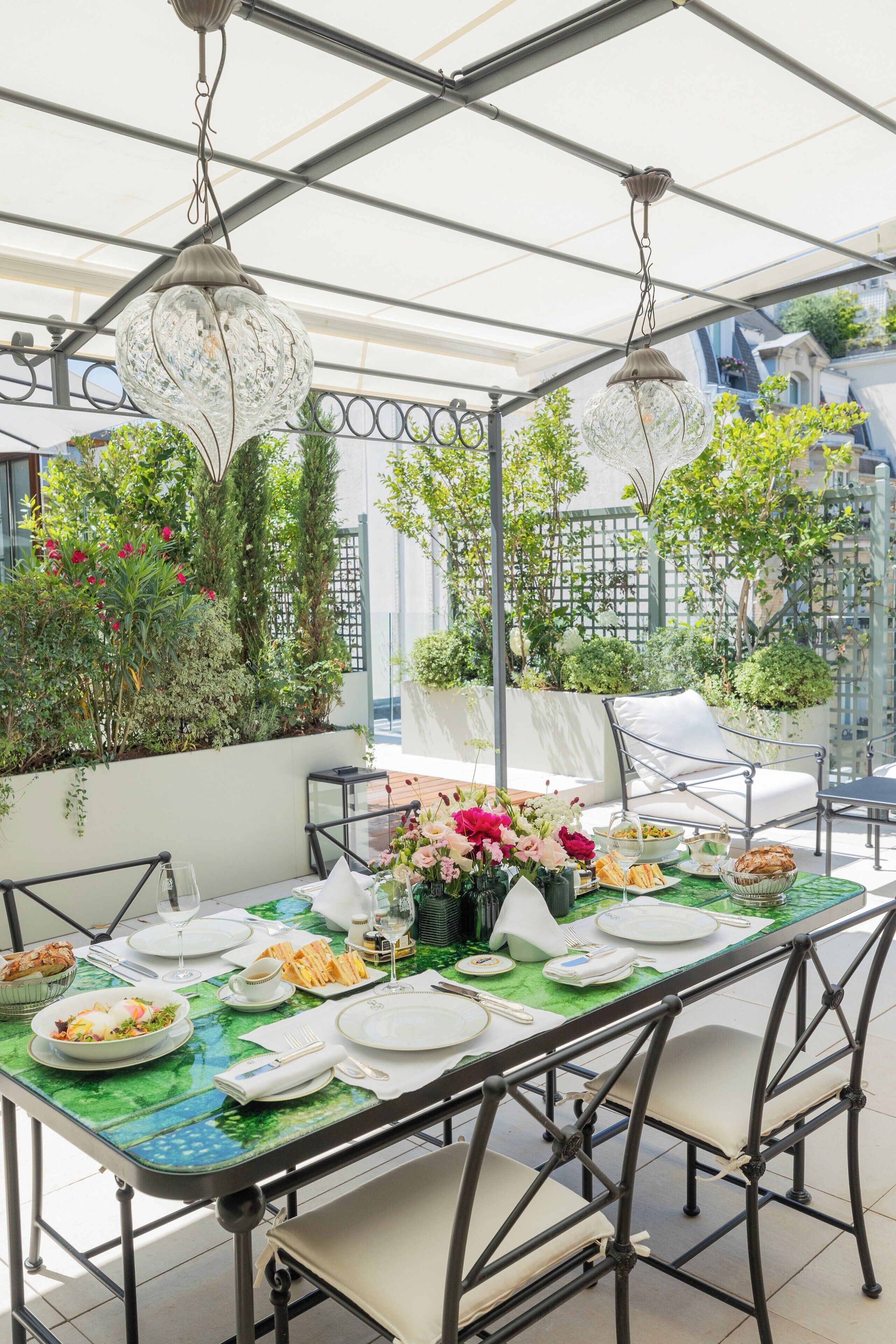 A dining area with a table set for a meal, surrounded by greenery and a view of buildings.
