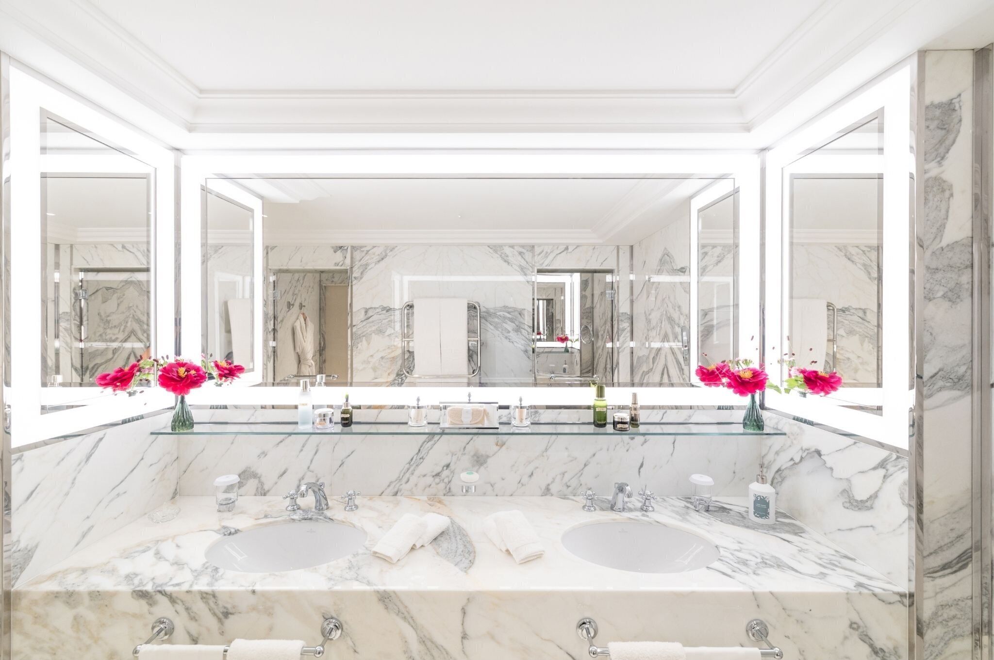 A luxurious bathroom with a large mirror, two sinks, marble countertops, and a glass shelf with toiletries.