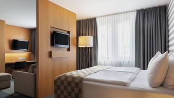 Quarto de hotel com uma cama, uma escrivaninha com cadeira, televisão e luminária.