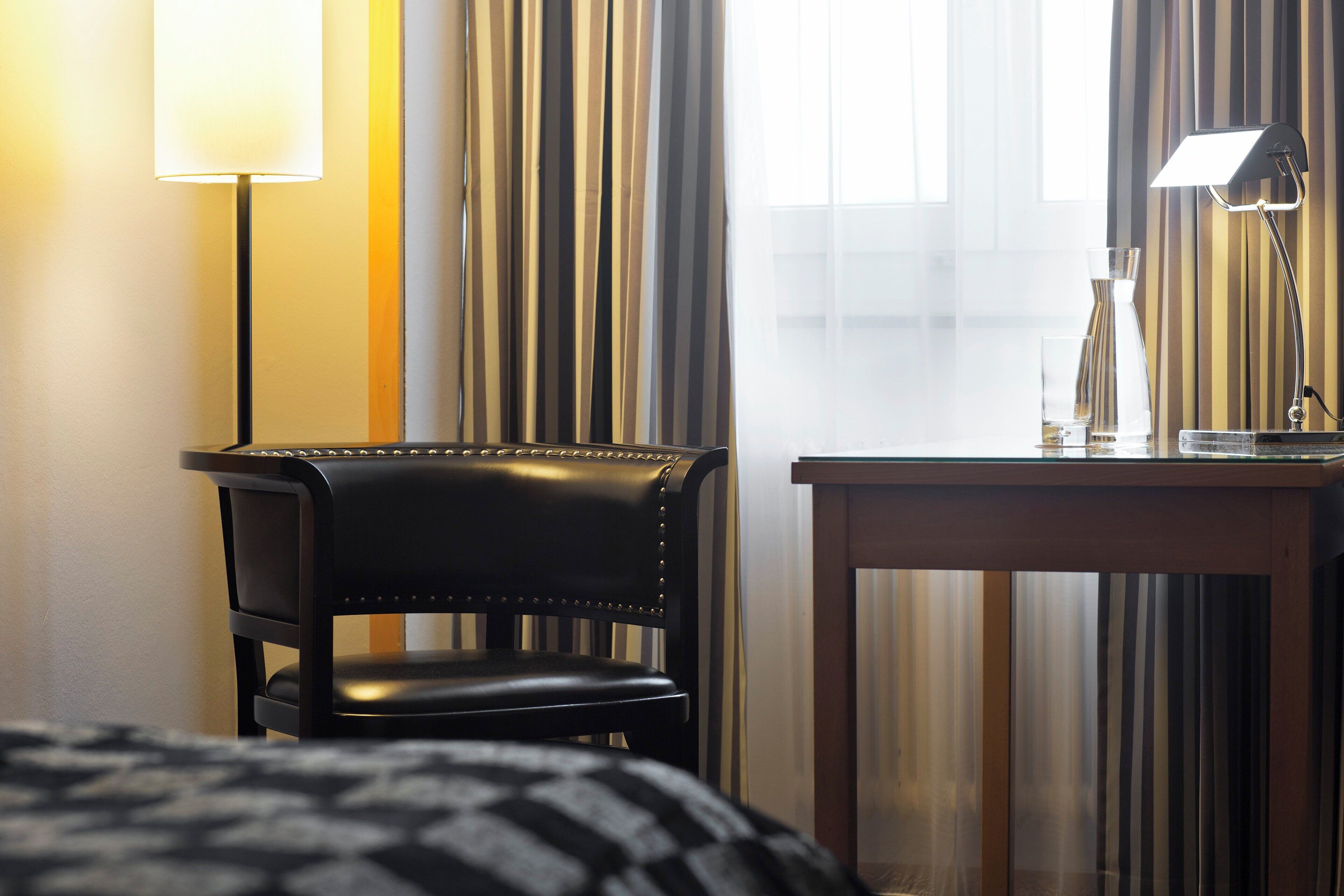 A hotel room with a bed, a chair, a table with a lamp, and a window with curtains.