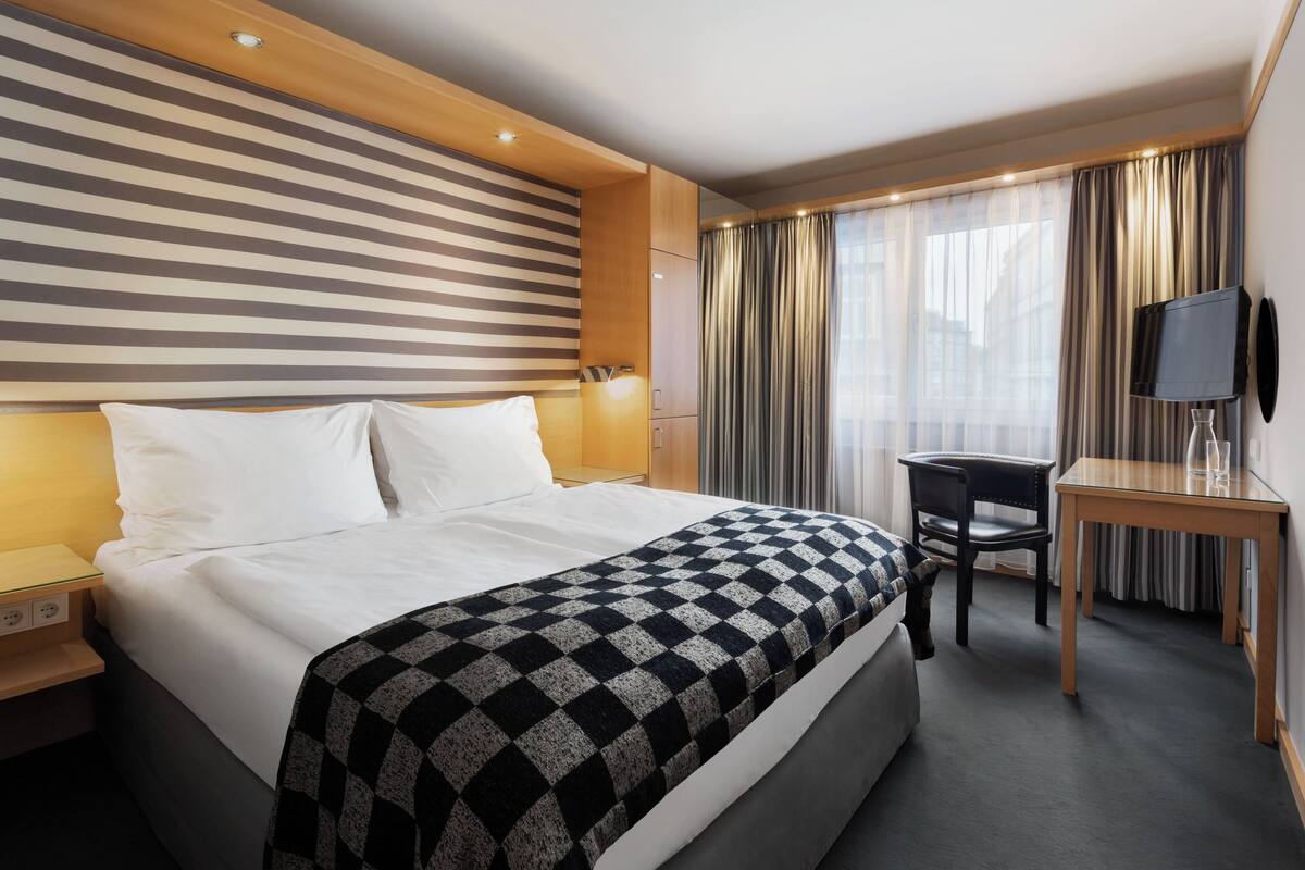 Une chambre d’hôtel avec un grand lit, un bureau avec une chaise, une télévision et une fenêtre avec des rideaux.
