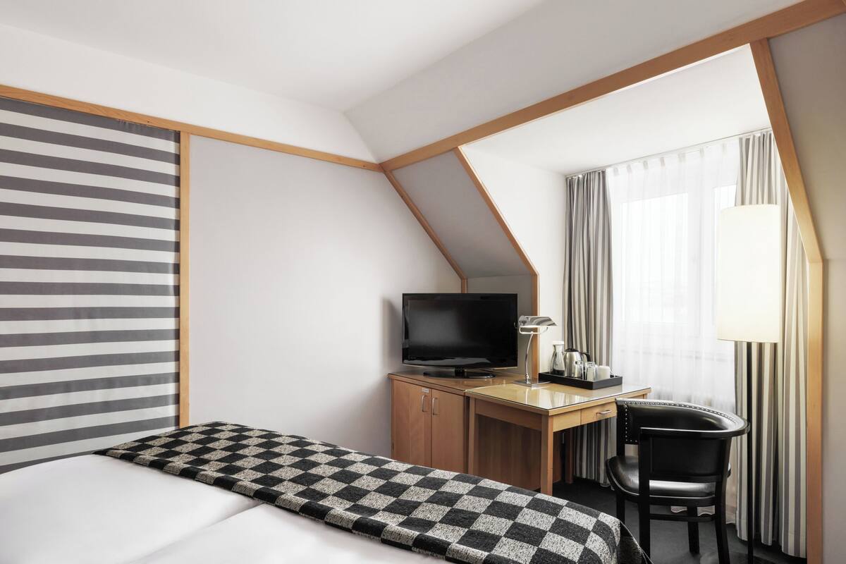 Une chambre d’hôtel avec un lit, un bureau avec une télévision, une chaise et une fenêtre avec des rideaux.