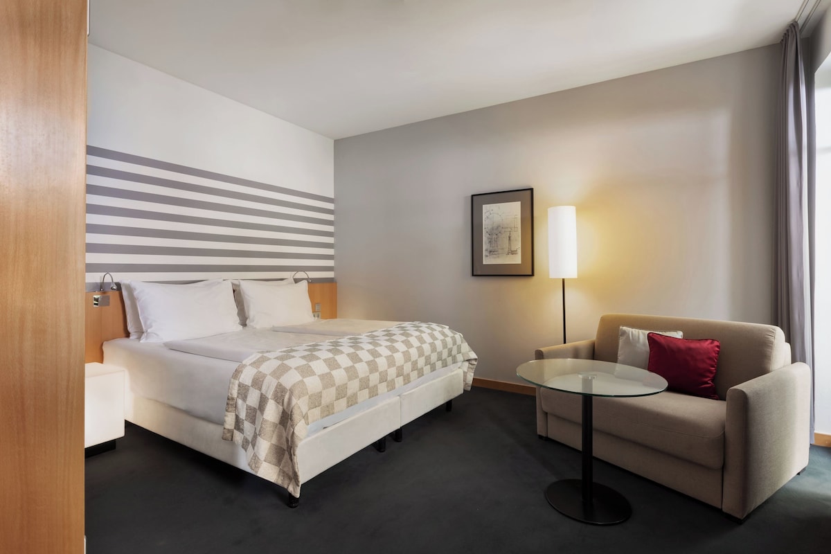 A hotel room with a bed, a sofa, a small table, a lamp, and a framed picture on the wall.