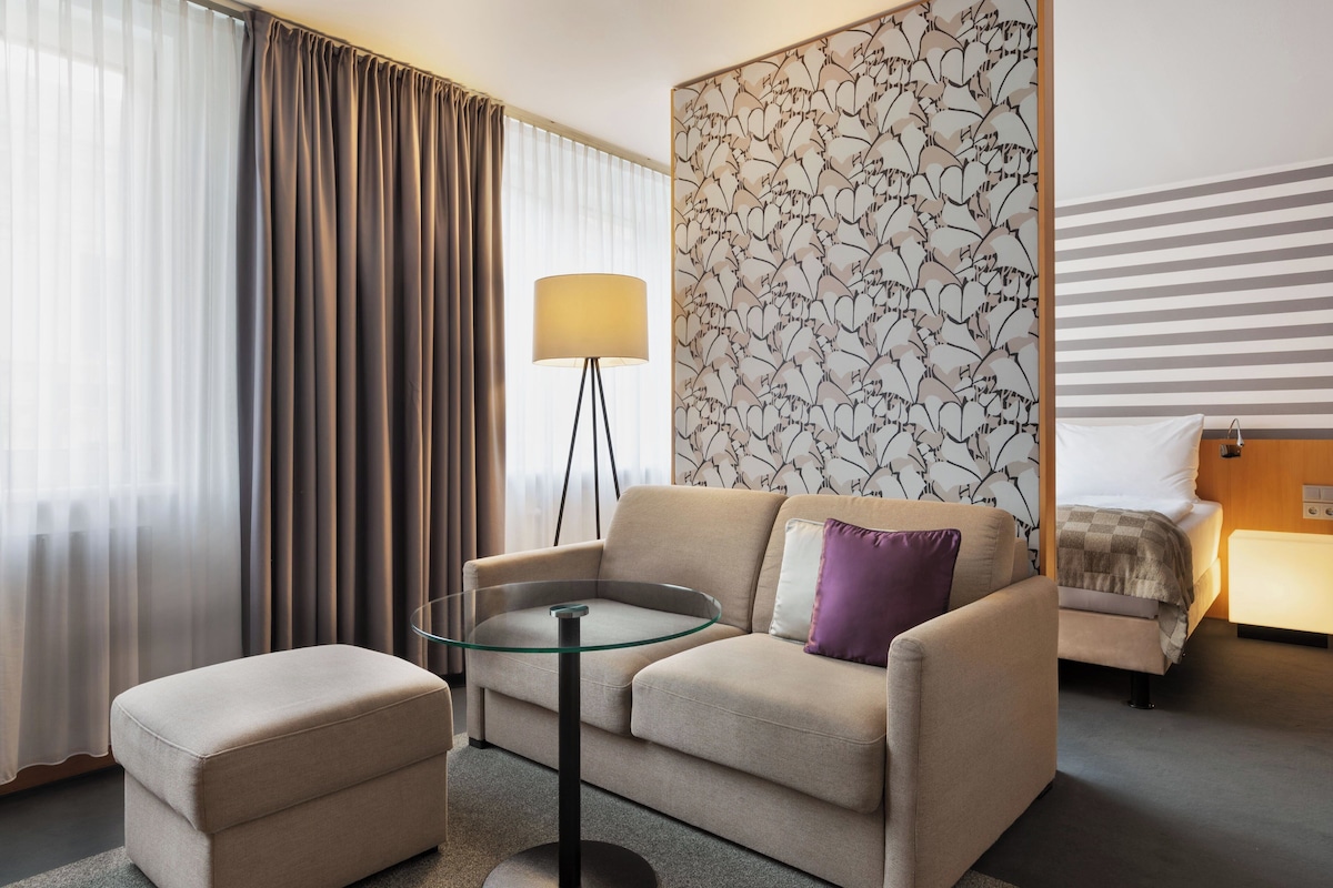 A hotel room with a sofa, ottoman, glass table, lamp, and a bed with a headboard.