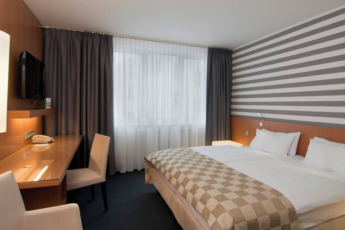 Une chambre d’hôtel comprenant un lit, un bureau avec une chaise, une télévision et une fenêtre avec des rideaux.