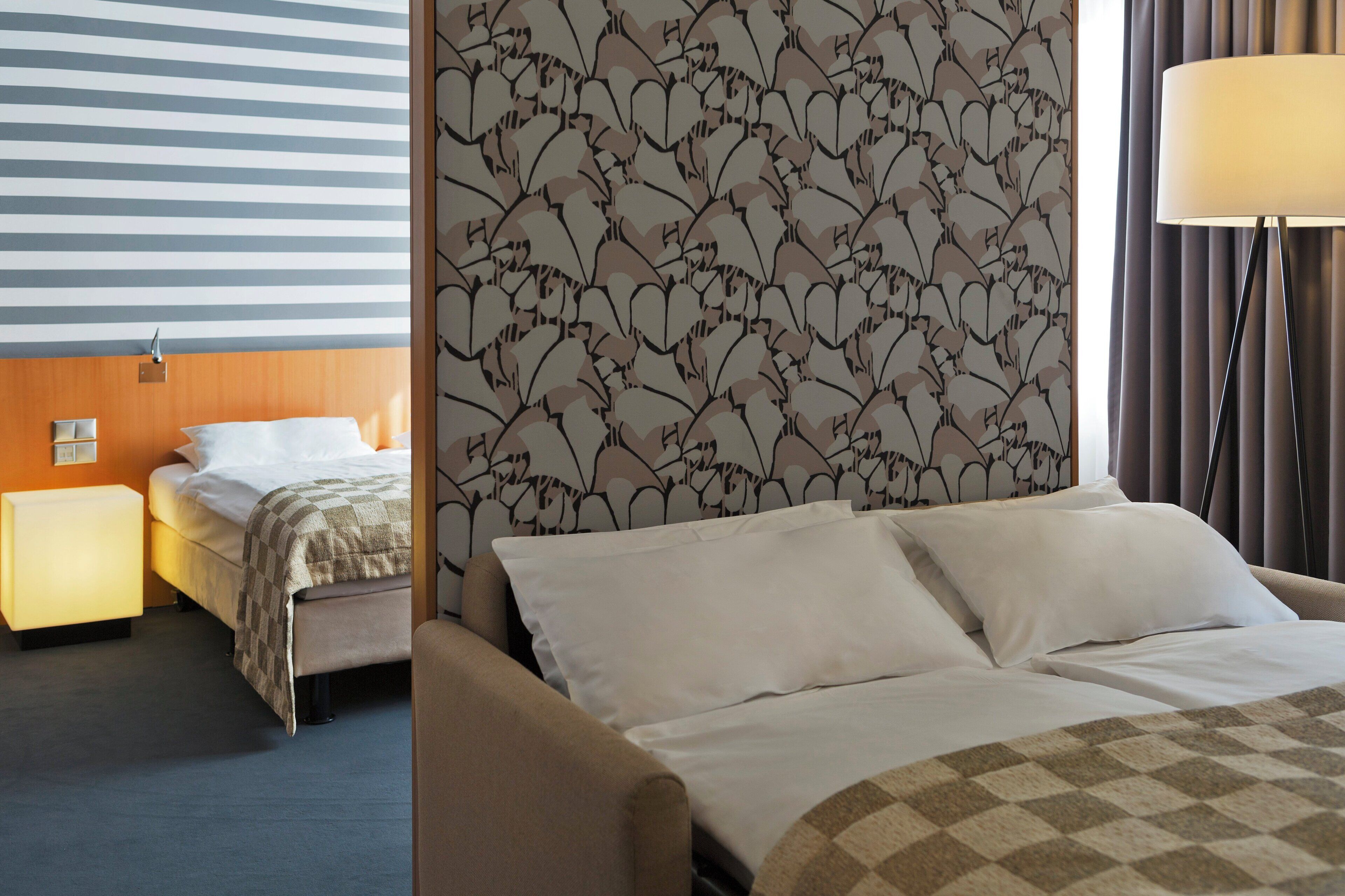 Una habitación de hotel con pared a rayas, un cabecero floral, una cama con ropa blanca, una lámpara de noche y una mesita de noche.