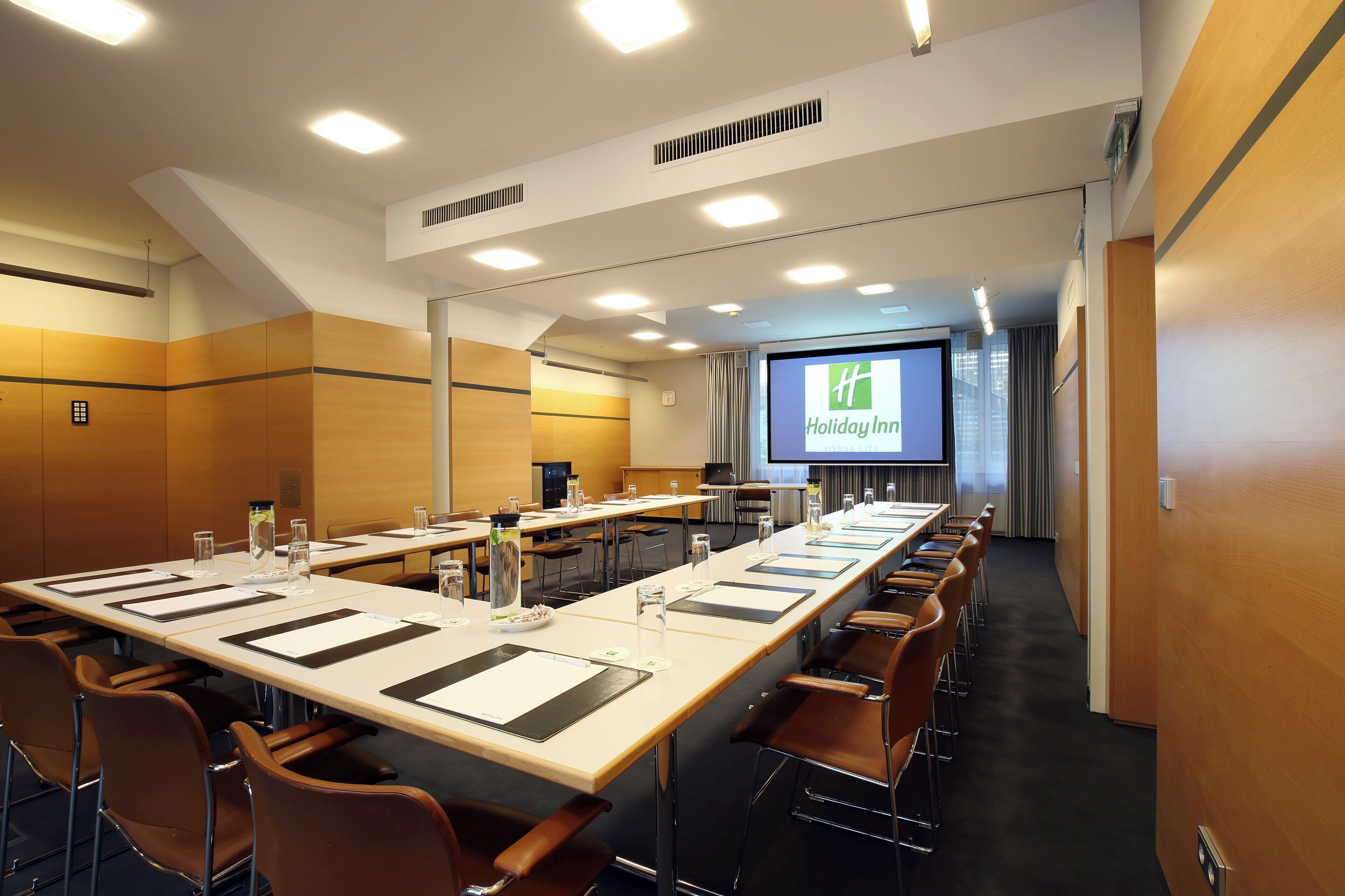 A conference room with a long table, chairs, and a screen displaying 'Holiday Inn'.