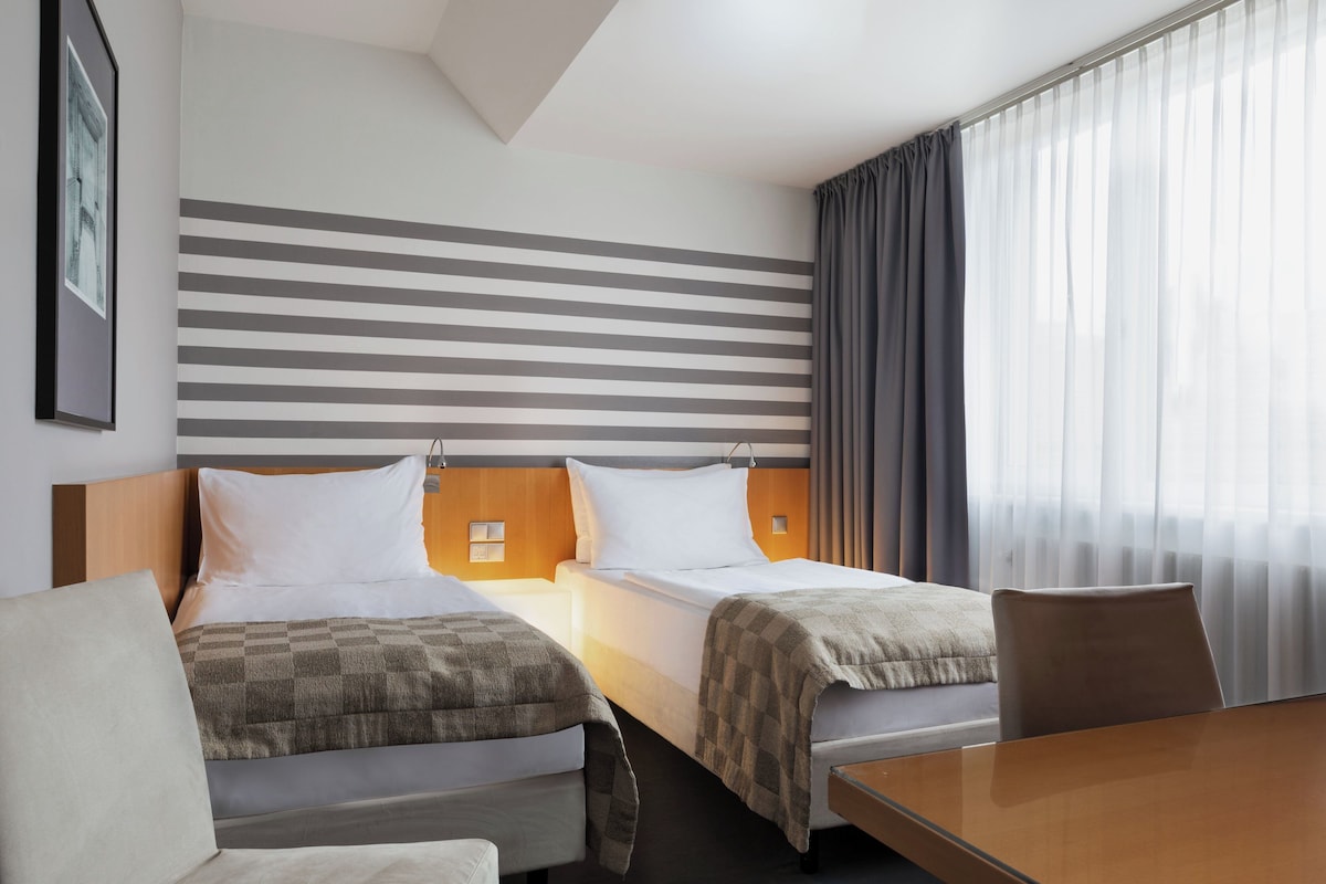 A hotel room with two beds, a wooden headboard, a large window with sheer curtains, and a wooden table.