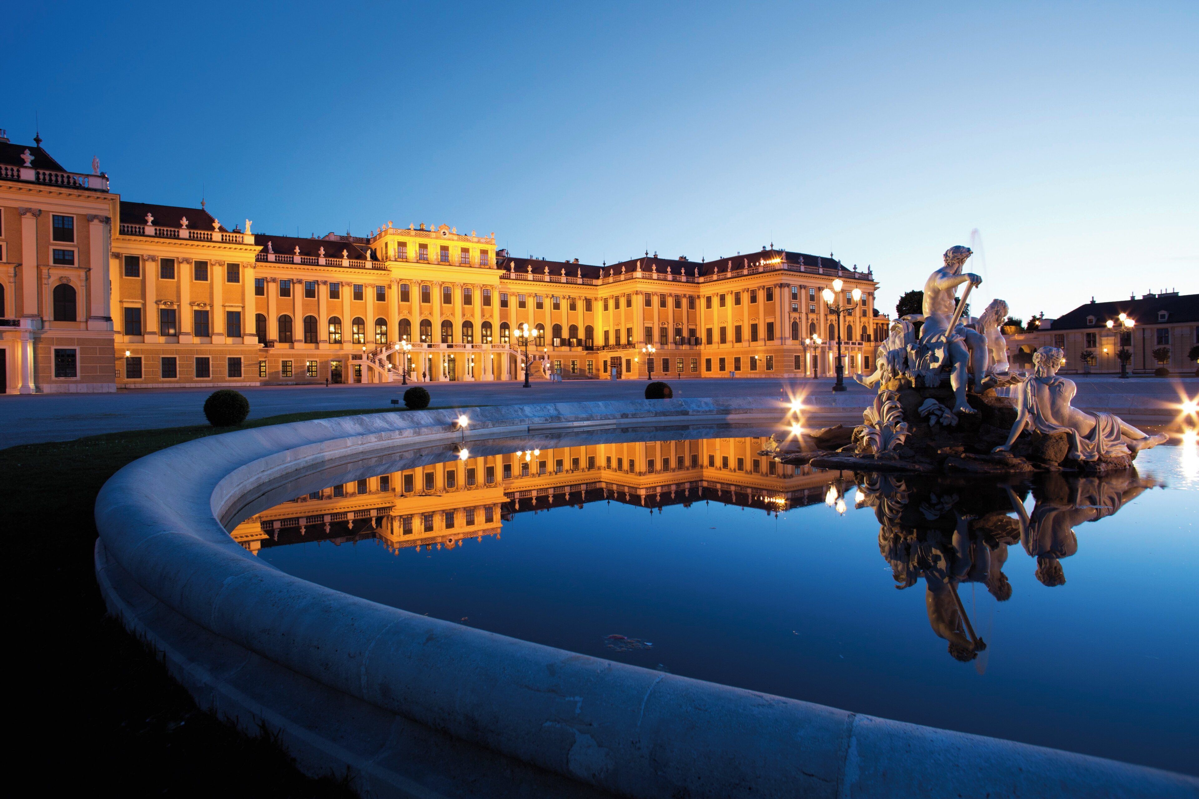 A grand building with classical architecture, a reflective water fountain, and a clear sky.