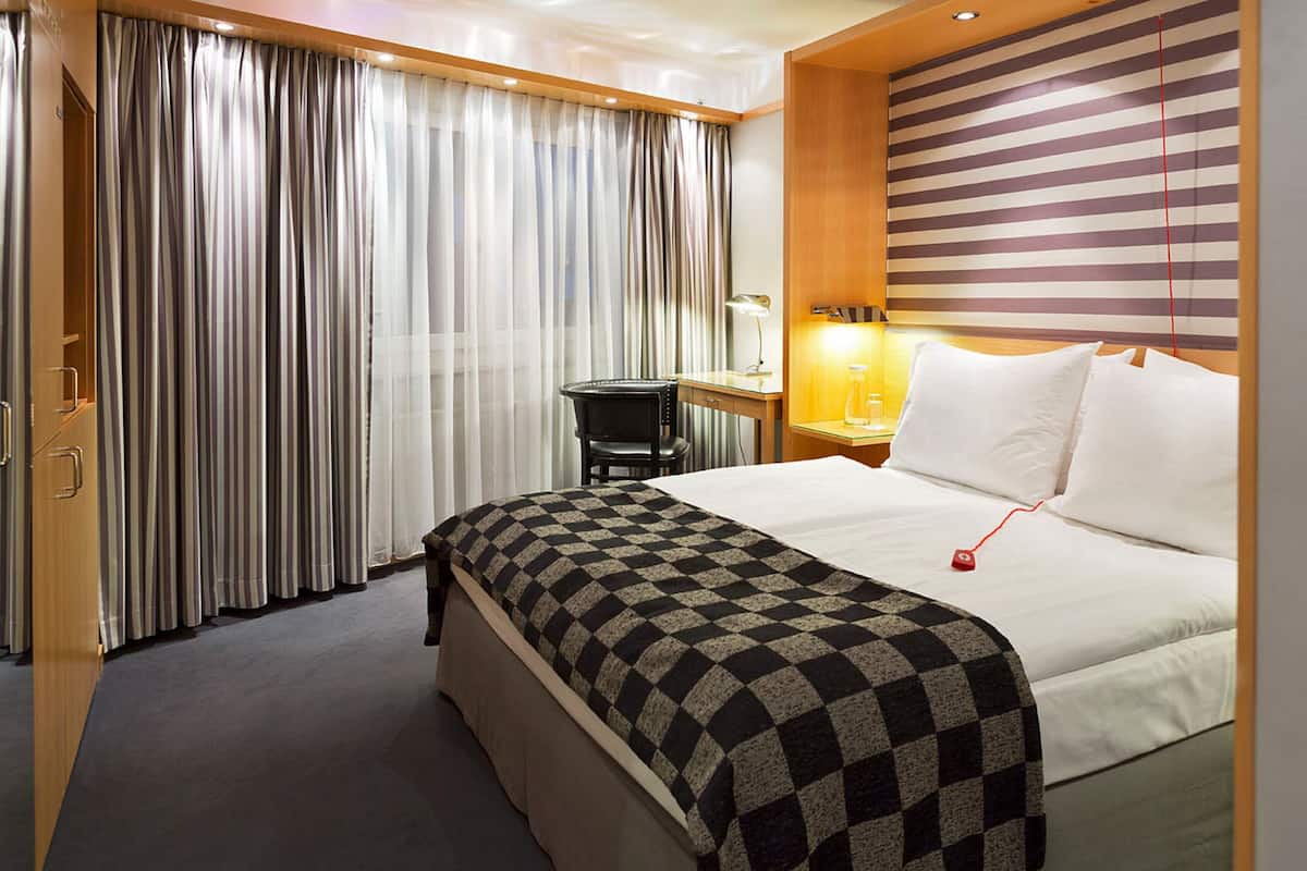 Une chambre d’hôtel avec un lit, un bureau, une chaise, une lampe et des rideaux.