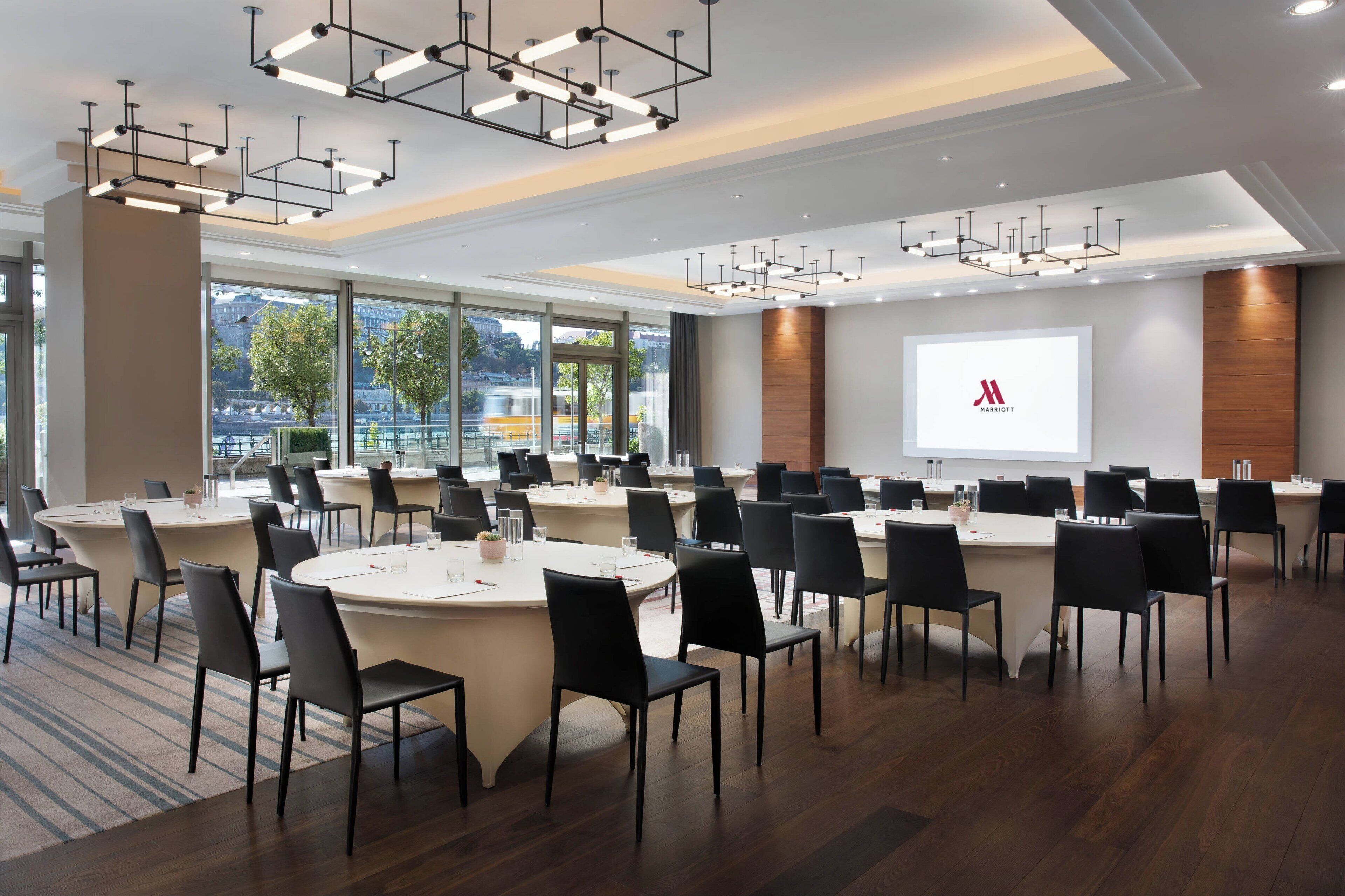 A conference room with multiple tables and chairs, large windows with a view of the city, and a presentation screen with the Marriott logo.