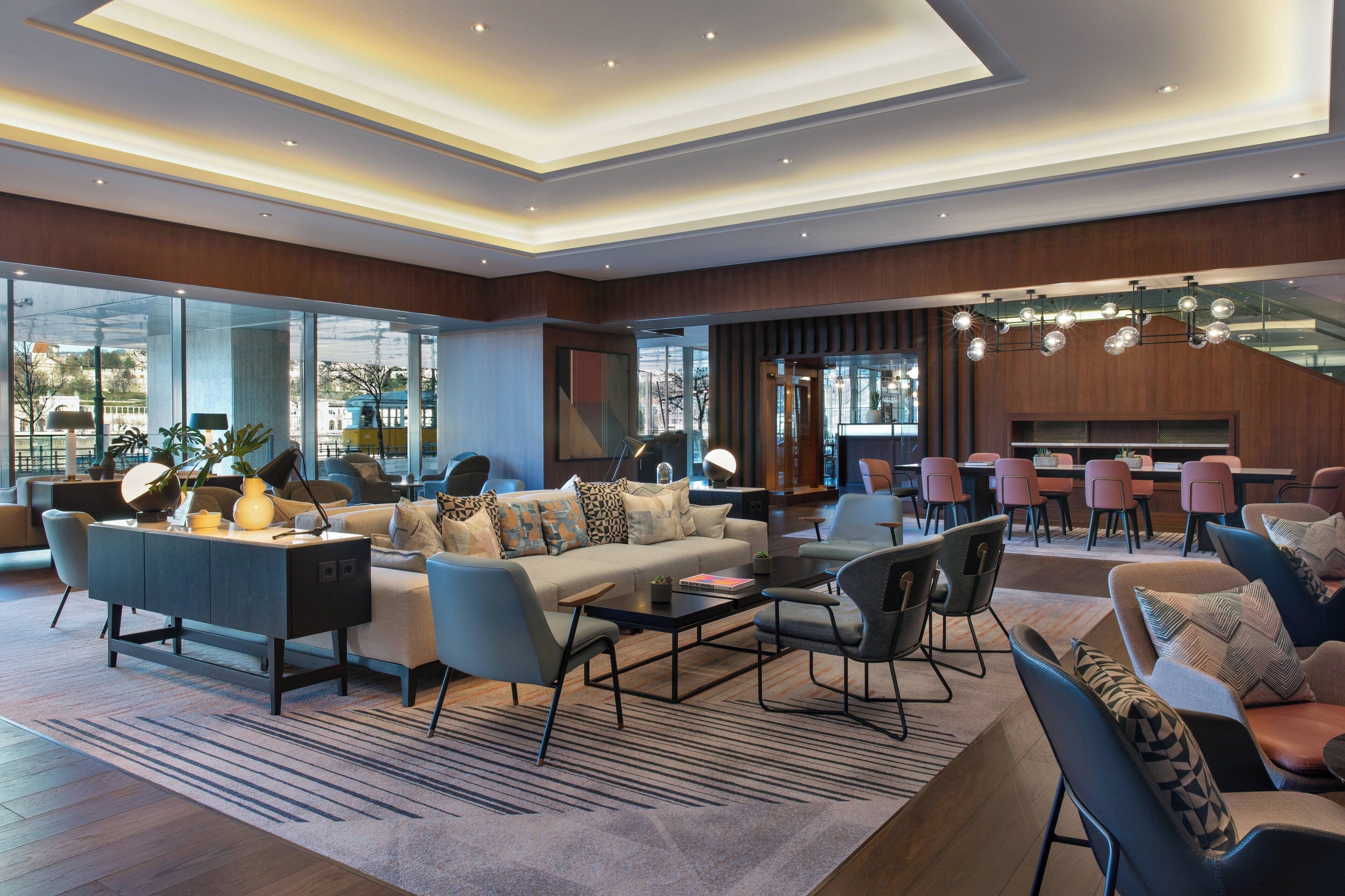 A modern hotel lobby with a sofa set, a coffee table, and a dining area with pink chairs and a wooden table.