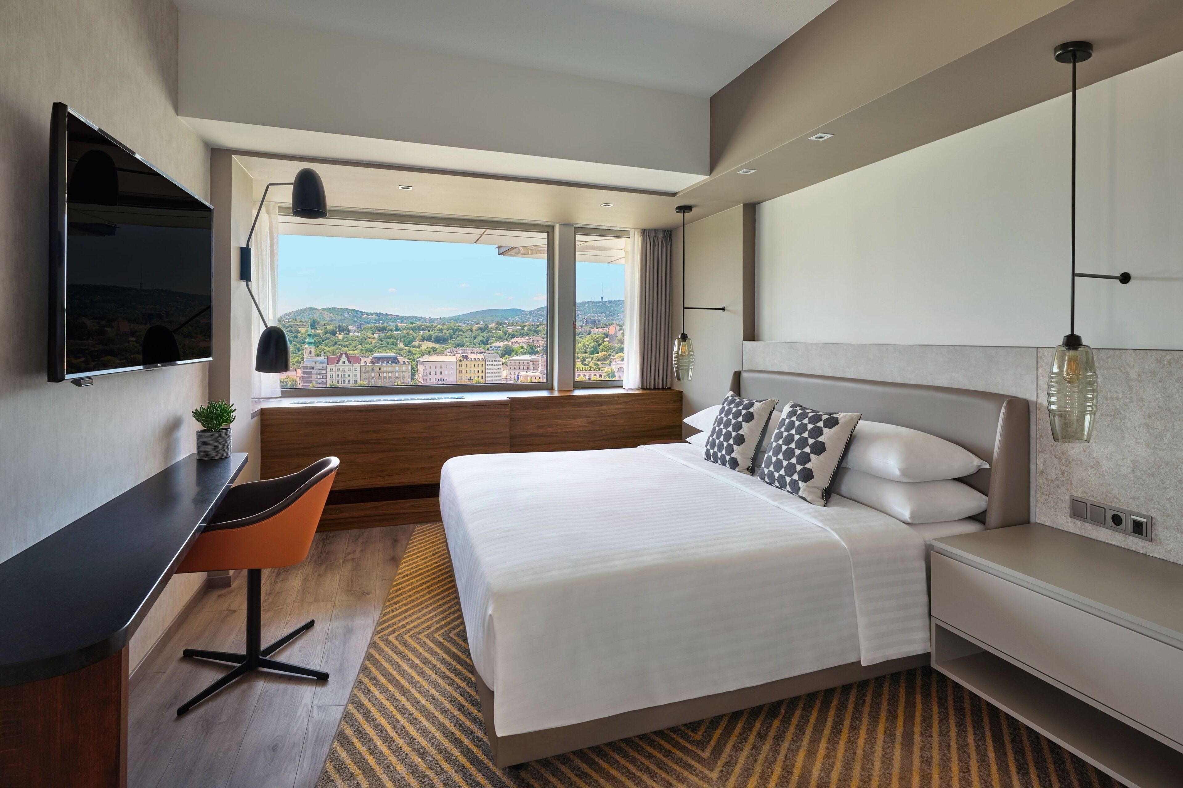 A modern hotel room with a large bed, a desk with a chair, a TV mounted on the wall, and a view of the cityscape through large windows.