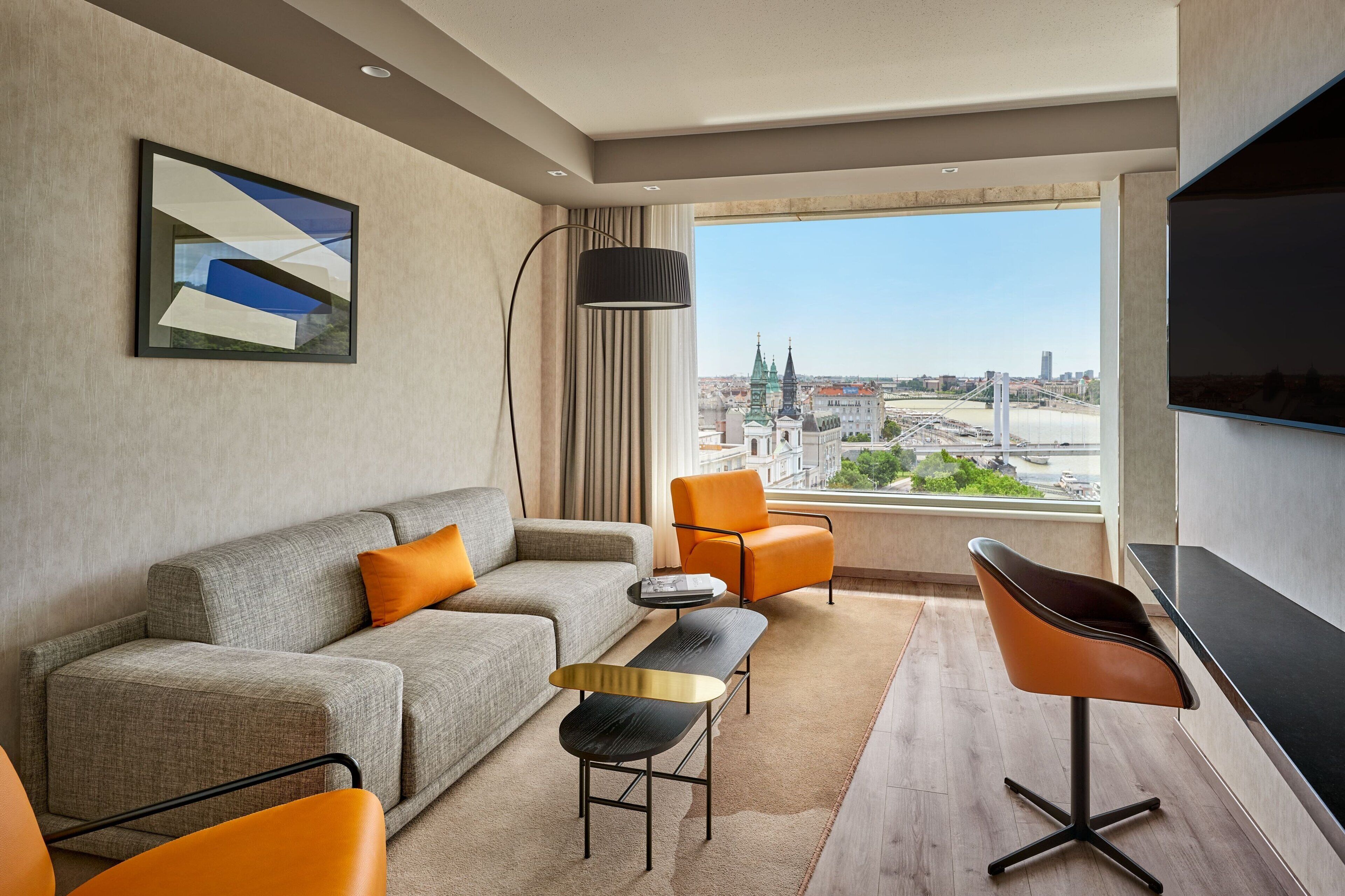 A modern living room with a sofa, chairs, a coffee table, and a TV. A large window offers a view of a cityscape.
