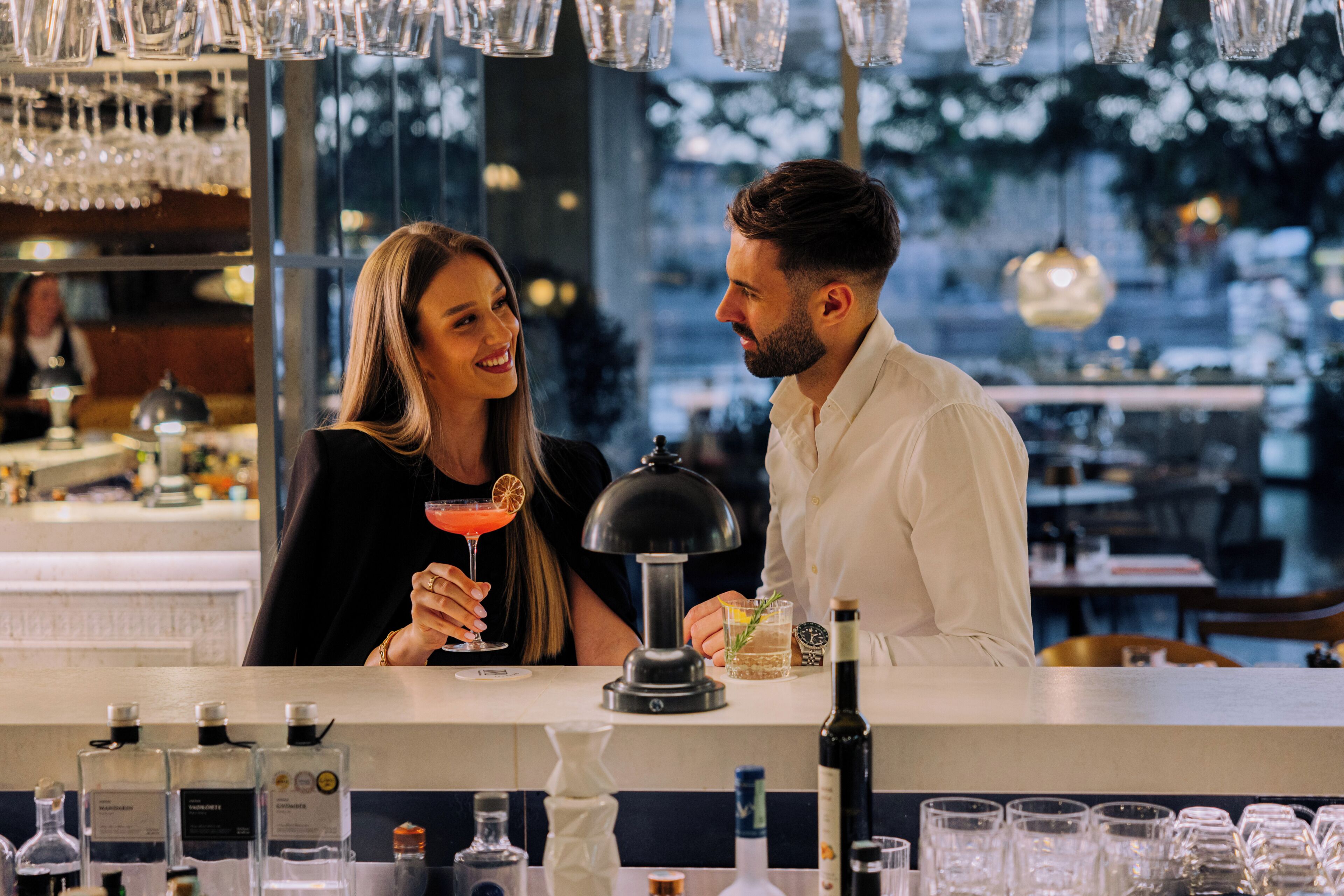 Una zona de bar con mostrador, vasos, botellas y una vista a la ciudad de fondo.