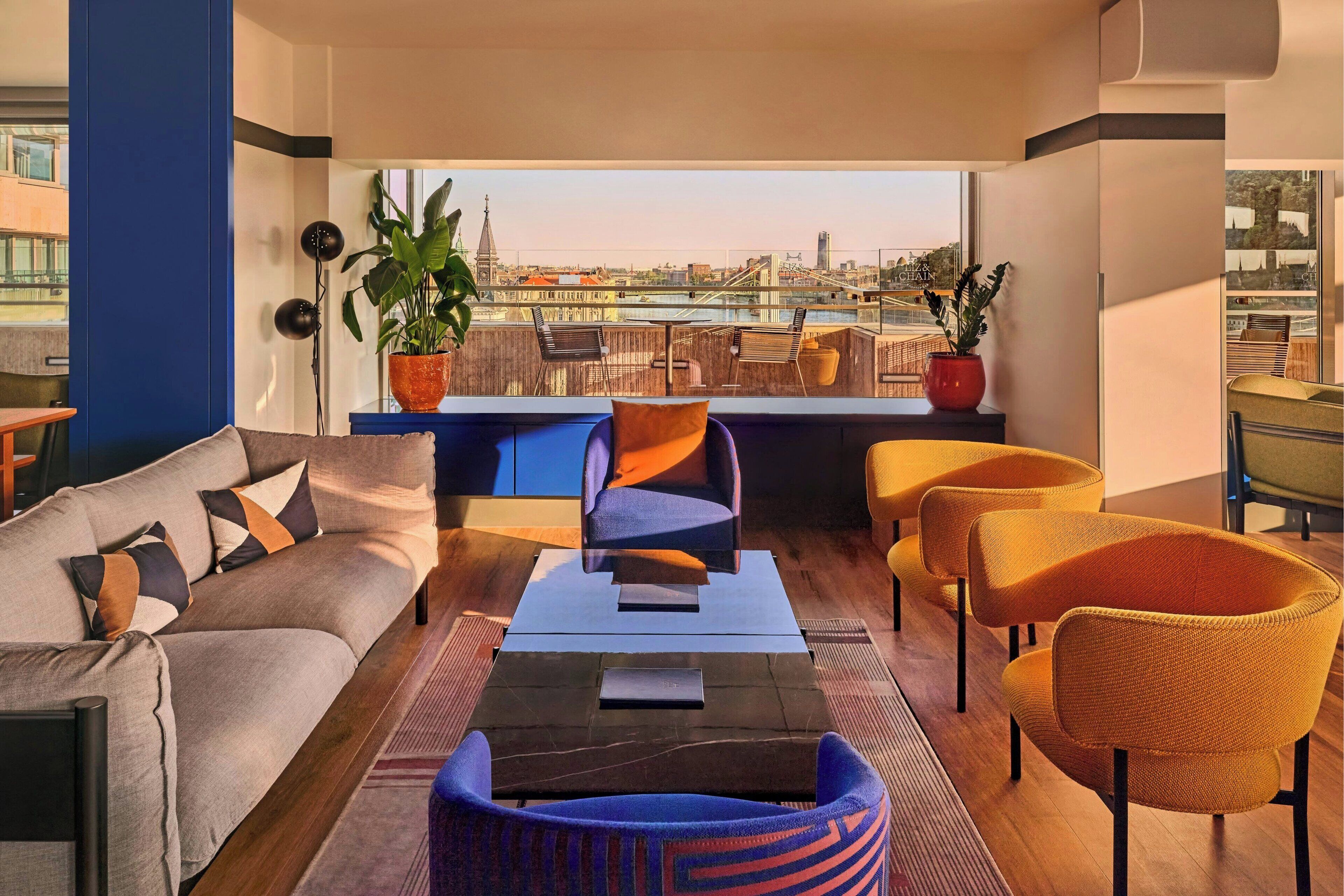 Una sala de estar moderna con un sofá, sillones y una mesa de centro. Hay plantas en macetas y una vista de la ciudad a través de amplias ventanas.