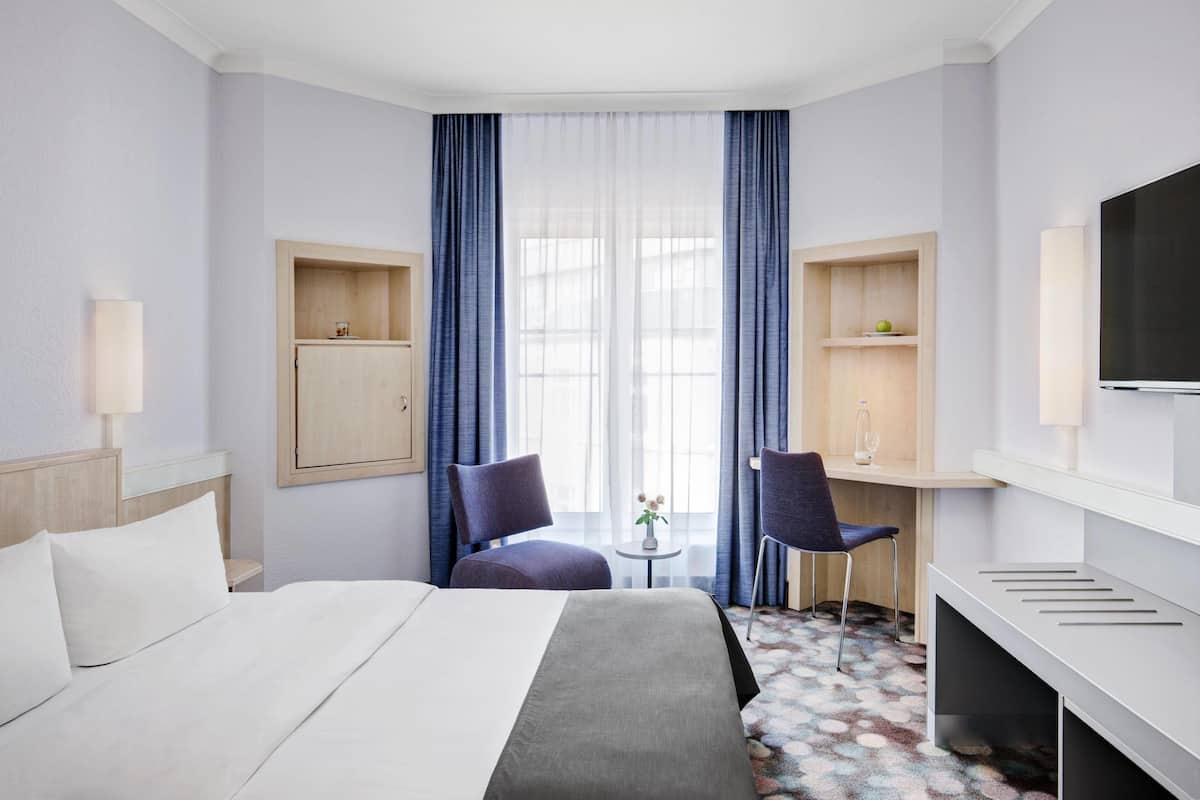 A hotel room with a bed, two chairs, a desk, a TV, and a window with curtains.