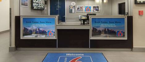 Zona de recepción con quiosco digital, tapete de bienvenida y carteles promocionales para una aplicación de motel.