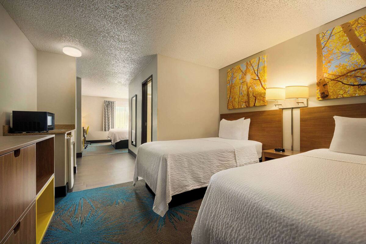 A hotel room with two beds, a desk with a television, a bathroom, and a hallway leading to another room.