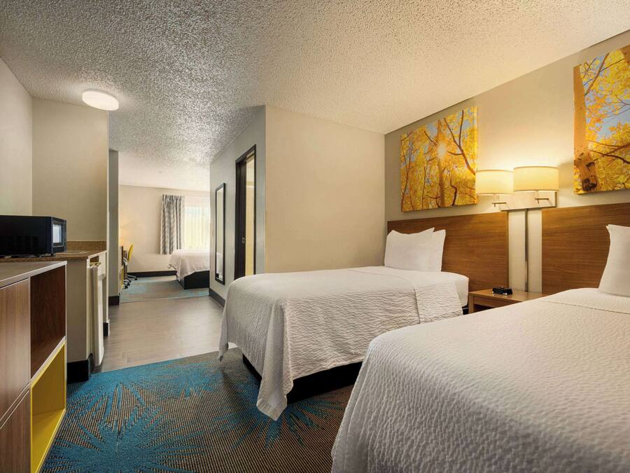 Habitación de hotel con dos camas, un escritorio con televisor, un baño y un pasillo que conduce a otra habitación.