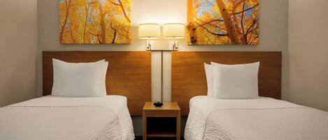 Habitación de hotel con dos camas, una mesita de noche con lámpara y dos cuadros colgados en la pared con árboles otoñales.