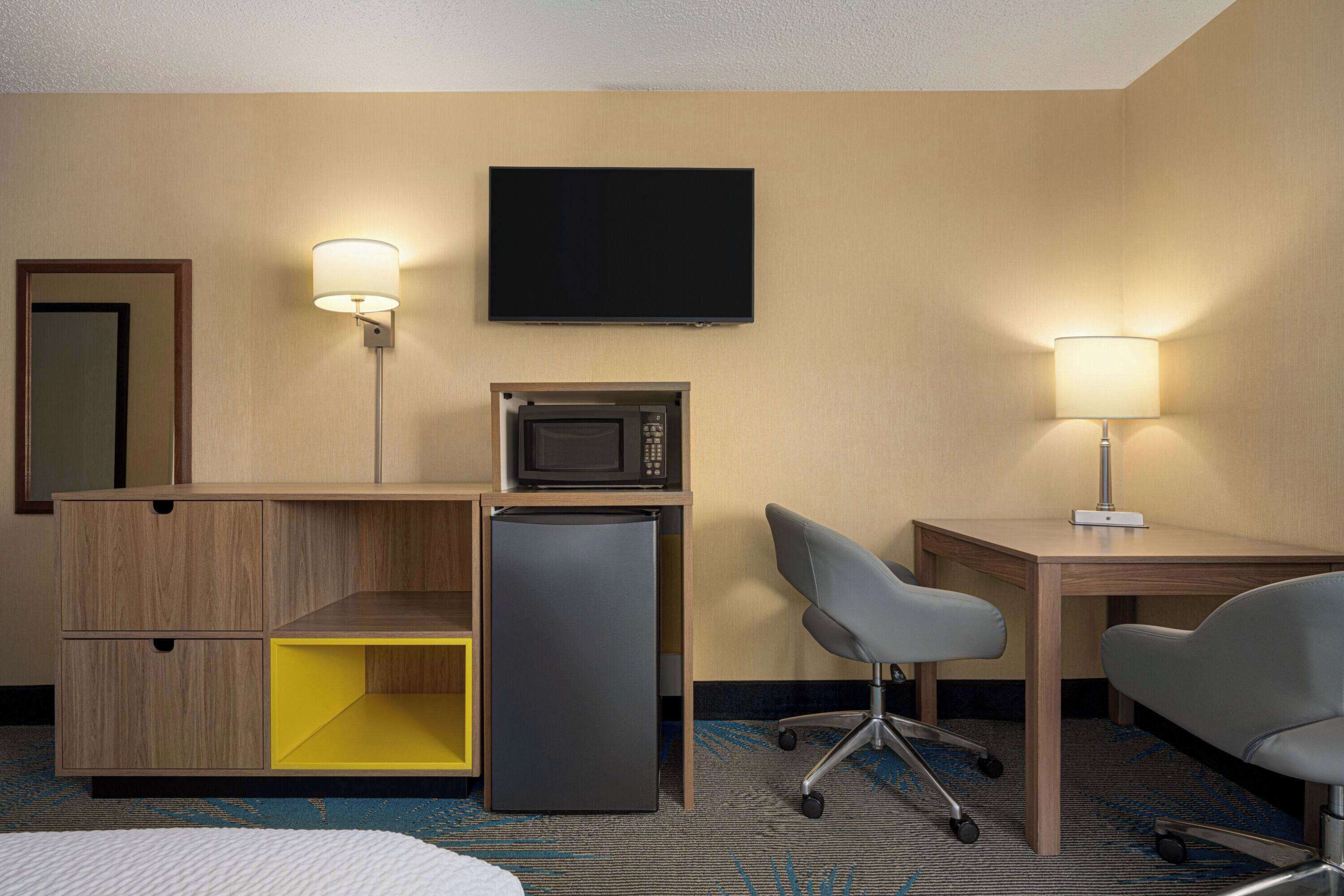 Ein Hotelzimmer mit einem Bett, einem Schreibtisch mit Computer, einer Mikrowelle, einem Kühlschrank, einem Fernseher und einer Lampe.