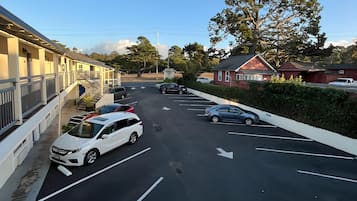 A parking lot with several cars, a white van, and a police car. There are buildings with balconies and a large tree in the background.