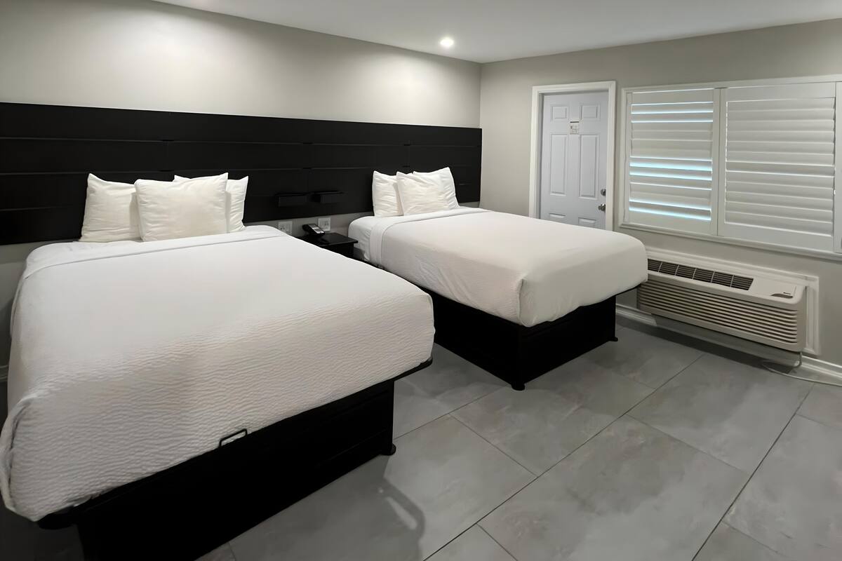 A hotel room with two beds, a black headboard, white bedding, a bedside table with a phone, a window with blinds, and a wall-mounted heater.