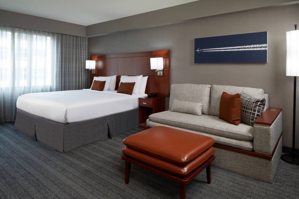 Ein Hotelzimmer mit einem großen Bett, einer Couch, einem Hocker, einem Nachttisch, einer Lampe und einem Wandbild.
