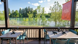 An outdoor seating area with tables and chairs, overlooking a lake with trees and a wooden deck. - Valley River Inn (Eugene)