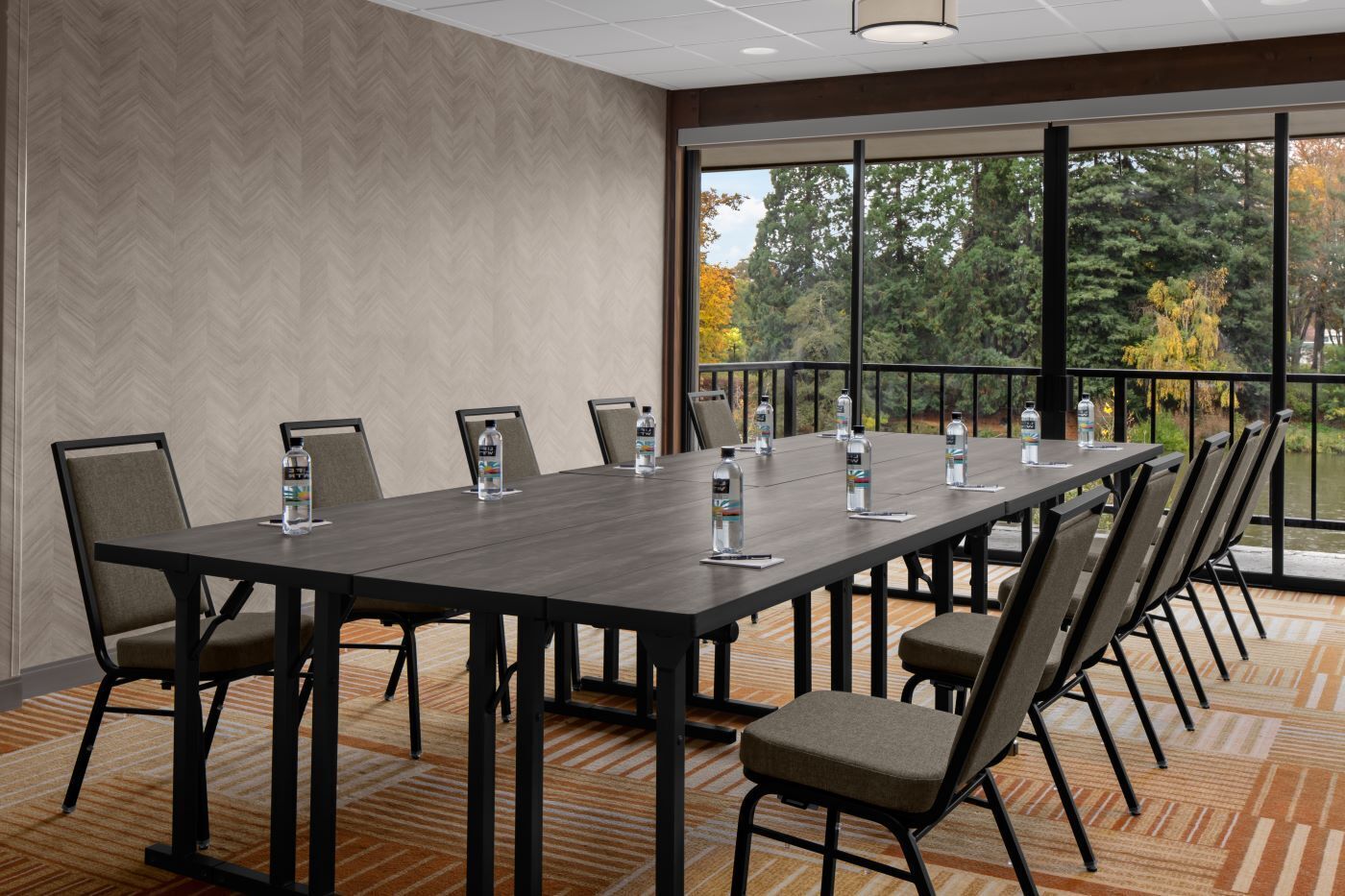 A conference room with a long table, chairs, water bottles, and a view of trees through large windows.