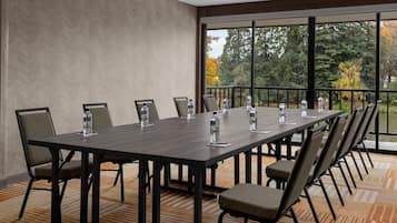 A conference room with a long table, chairs, water bottles, and a view of trees through large windows.