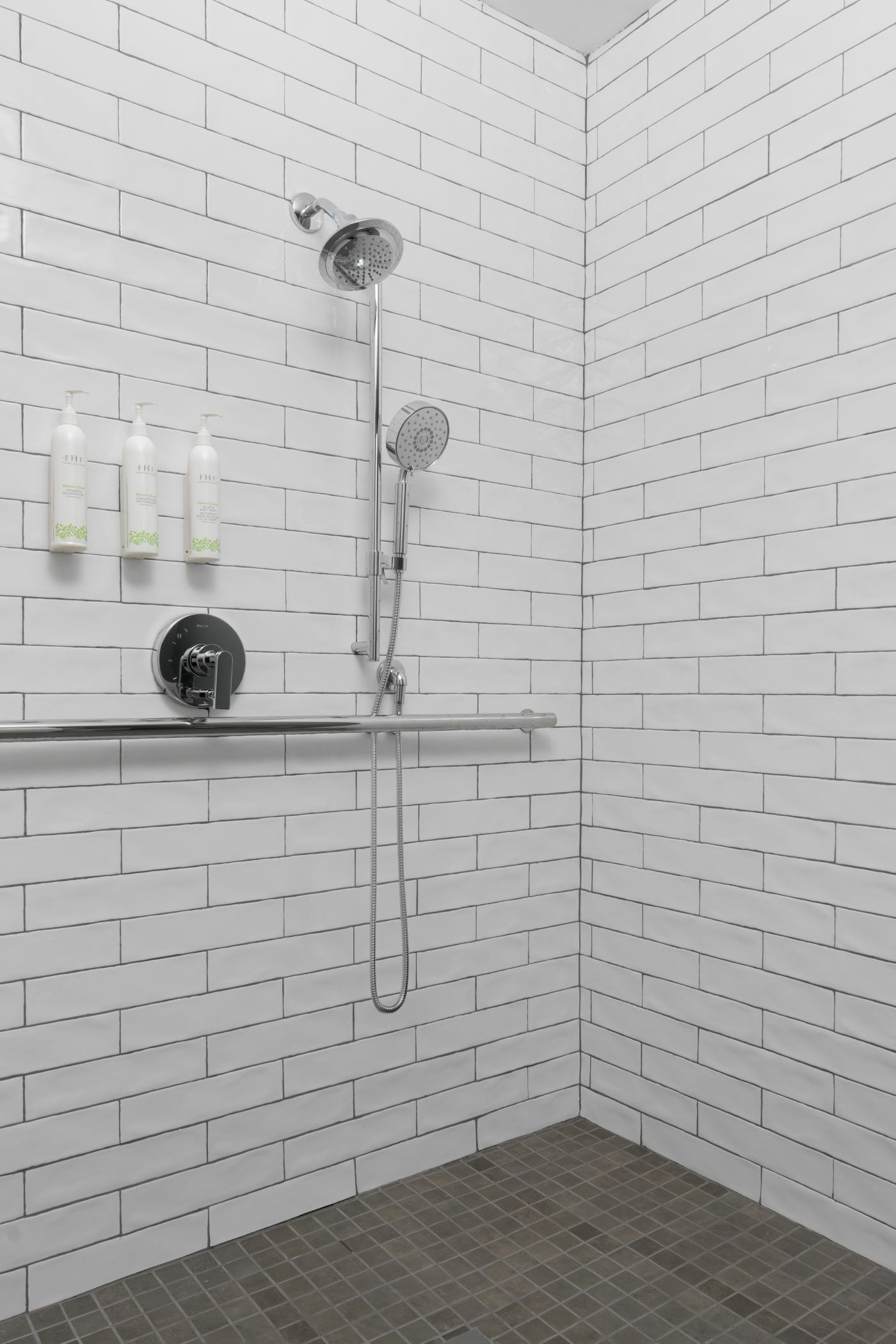 Un espace douche avec des murs et un sol recouverts de carreaux blancs, une douchette à main, un robinet mural et un distributeur de savon.