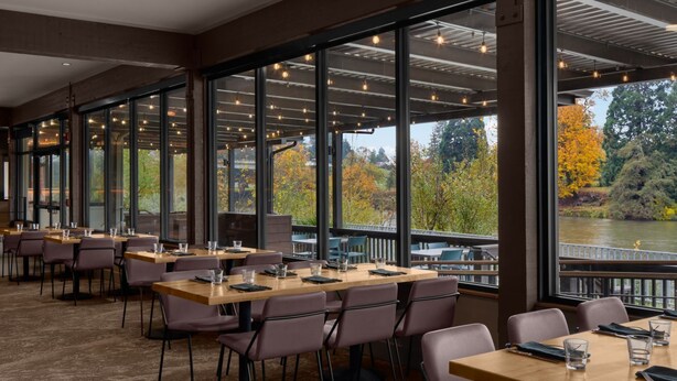 A restaurant with large windows offering views of a river and autumn trees. Tables are set with chairs and glasses.